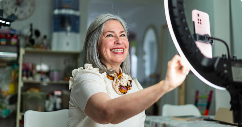 Image of an older woman smiling in front of a camera phone with a light ring fixed round it, as if filming for a social media post